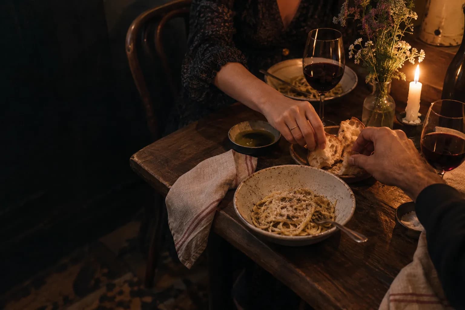 Two people sharing bread over pasta and wine at a candlelit table